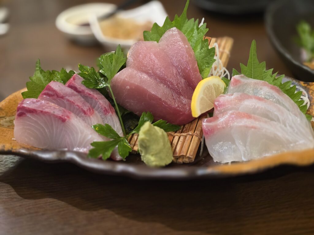 Assorted sashimi slices of tuna, white fish, and yellowtail arranged on a ceramic plate with shiso leaves, lemon wedge, and wasabi