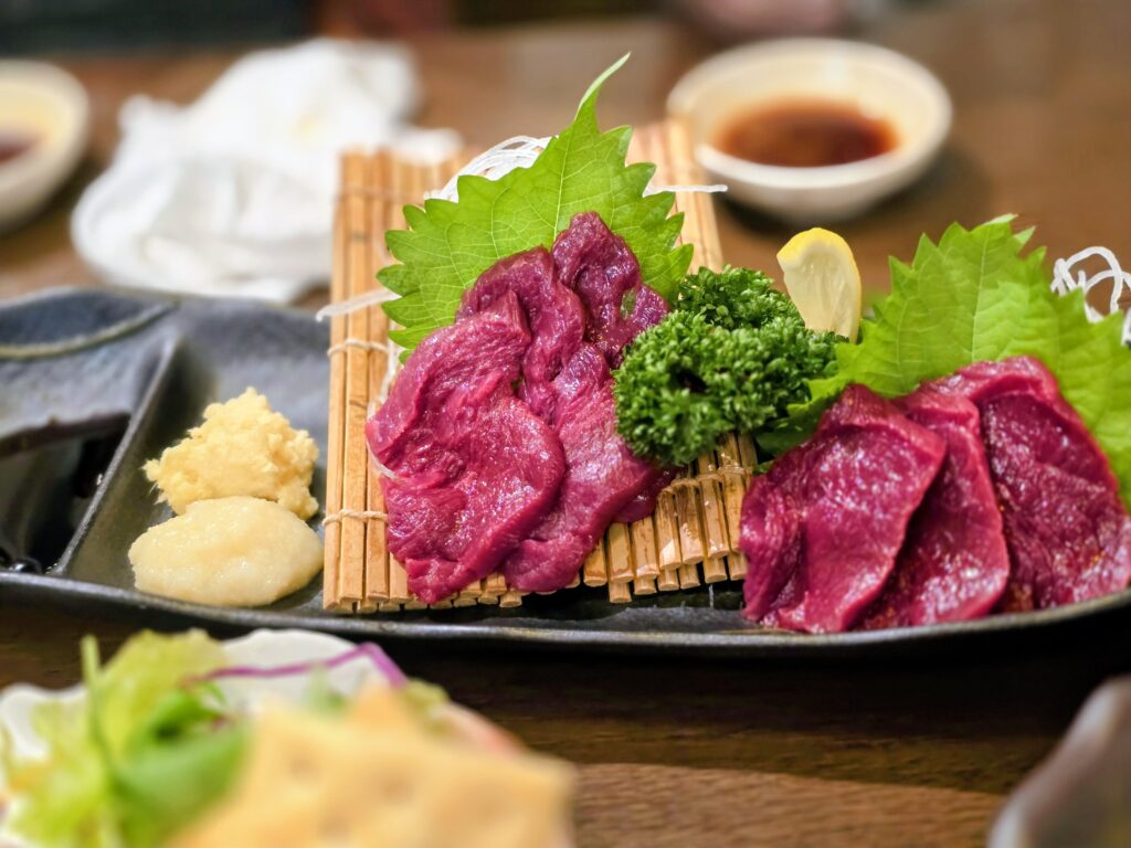 Plate of fresh horse meat sashimi served with ginger, garlic, and leafy garnish on a bamboo mat.
