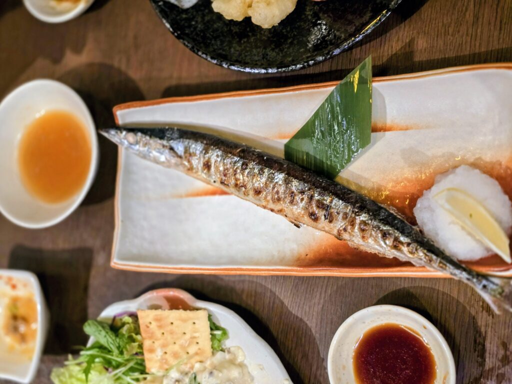 Whole grilled Pacific saury fish served on a rectangular plate with grated daikon and lemon wedge.