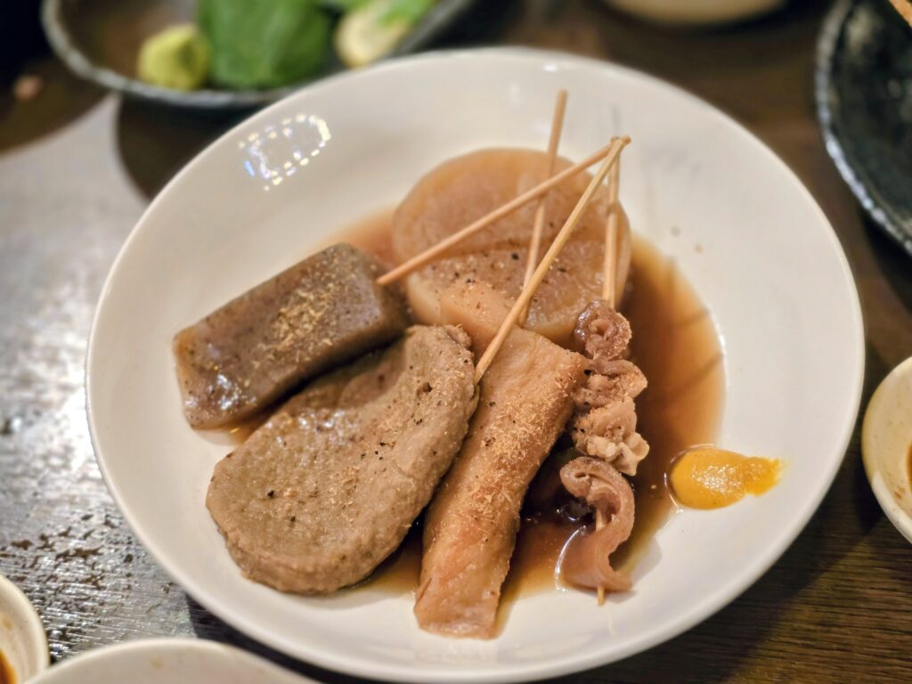 Bowl of simmered oden with radish, konjac, fish cakes, and skewered beef in a soy broth with mustard on the side.
