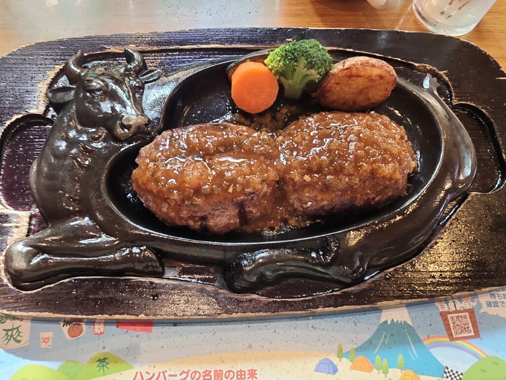 Juicy hamburg steak served sizzling on a cow-shaped iron plate with onion sauce, carrot, broccoli, and potato.