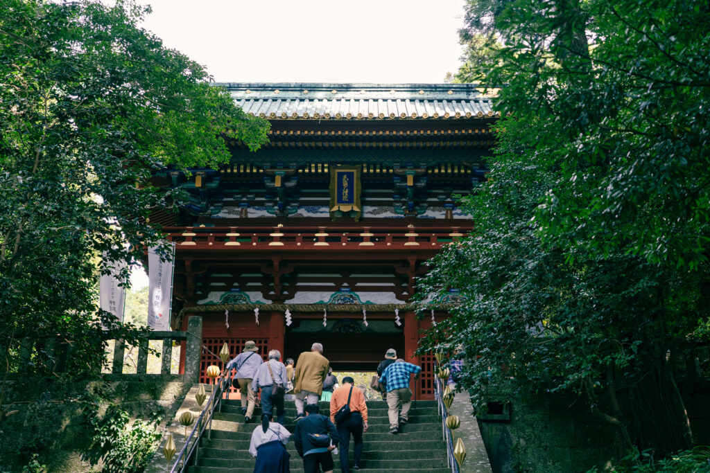 Group of visitors climbing stone steps toward the red and gold gate of Nihondaira’s Kunōzan Tōshōgū Shrine, framed by green trees.