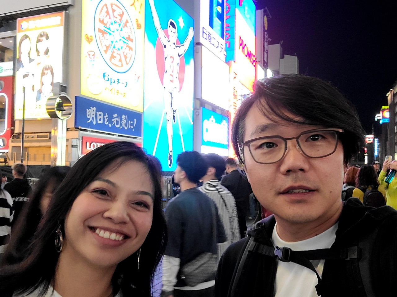 A smiling couple posing in front of the iconic Glico Running Man sign in Dotonbori, Osaka at night. The background glows with colorful neon lights and a lively crowd, capturing the energy of the city’s nightlife.