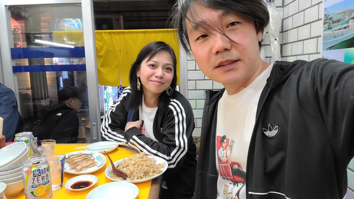 A couple enjoying dinner together at a cozy Japanese eatery. They are seated at a bright yellow table with plates of fried rice, gyoza, and canned drinks. The casual atmosphere and tiled walls create a warm, local diner feel.