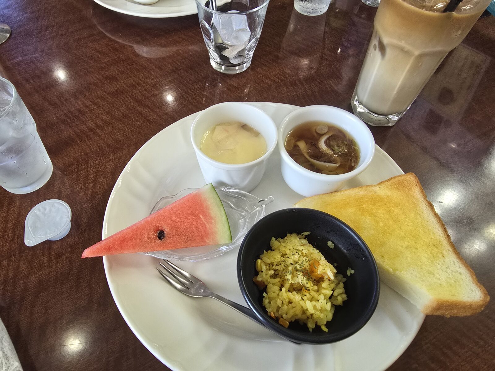 A generous Gifu morning set featuring toast, rice pilaf, chawanmushi egg custard, miso soup, and a slice of watermelon, served with iced coffee.