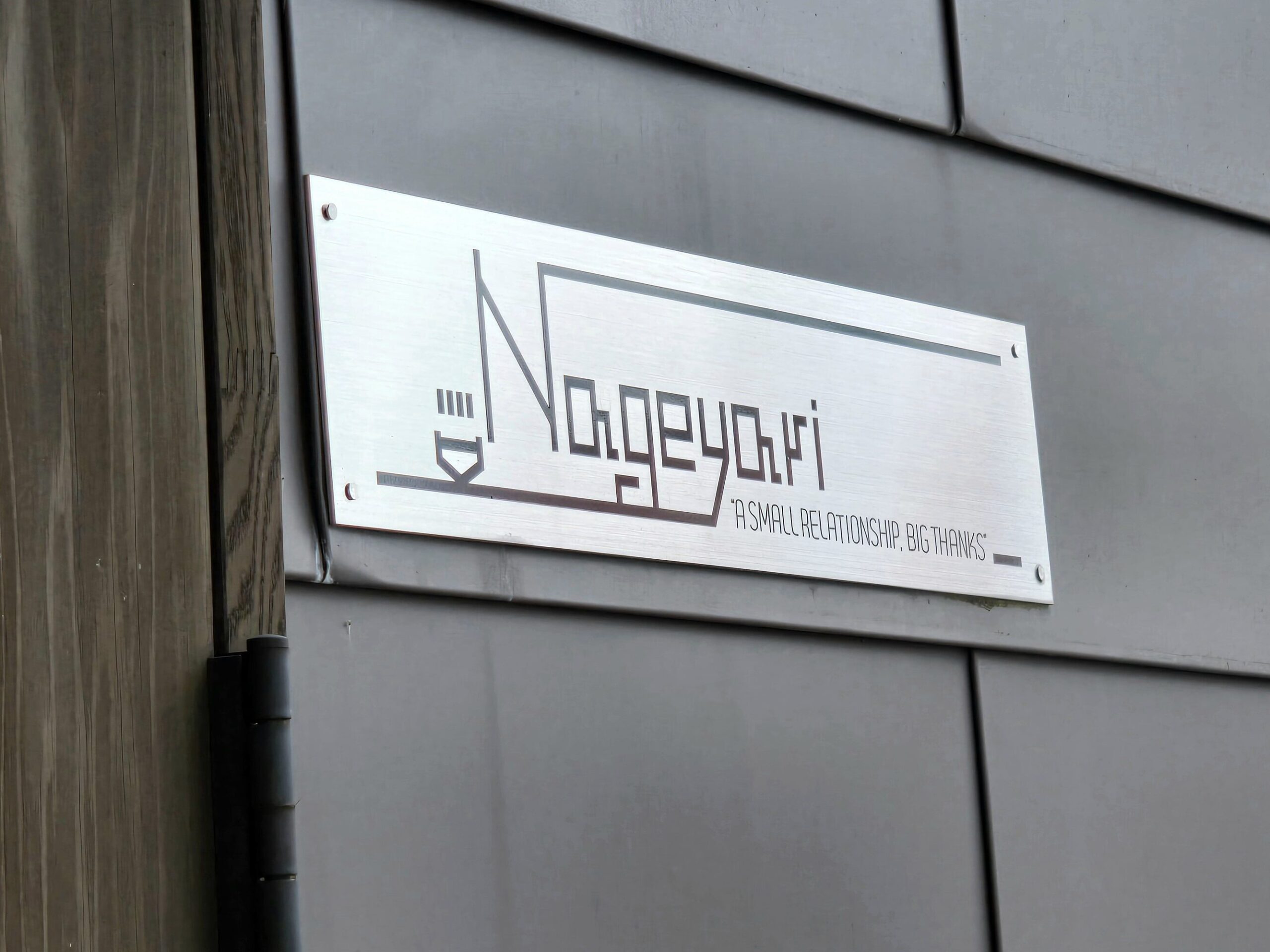 The minimalist metal sign of Nageyari ramen shop in Gifu City, Japan, engraved with its logo and the phrase “A small relationship; big thanks.