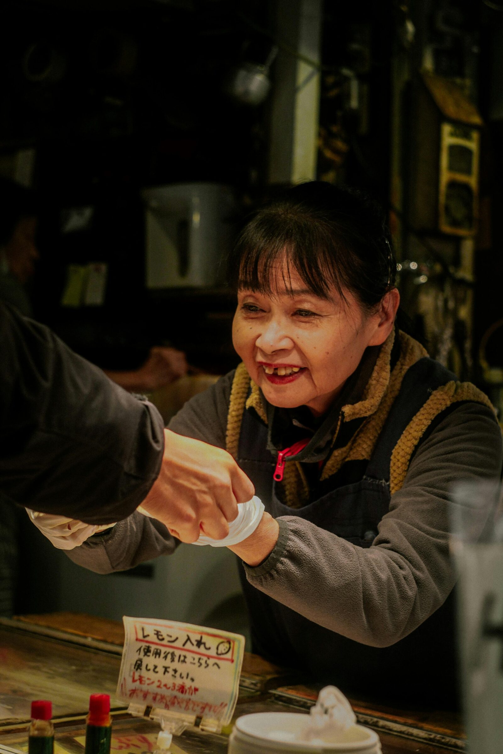 A smiling woman at a small Japanese food stall warmly handing an item to a customer, representing genuine friendliness and care in daily service.