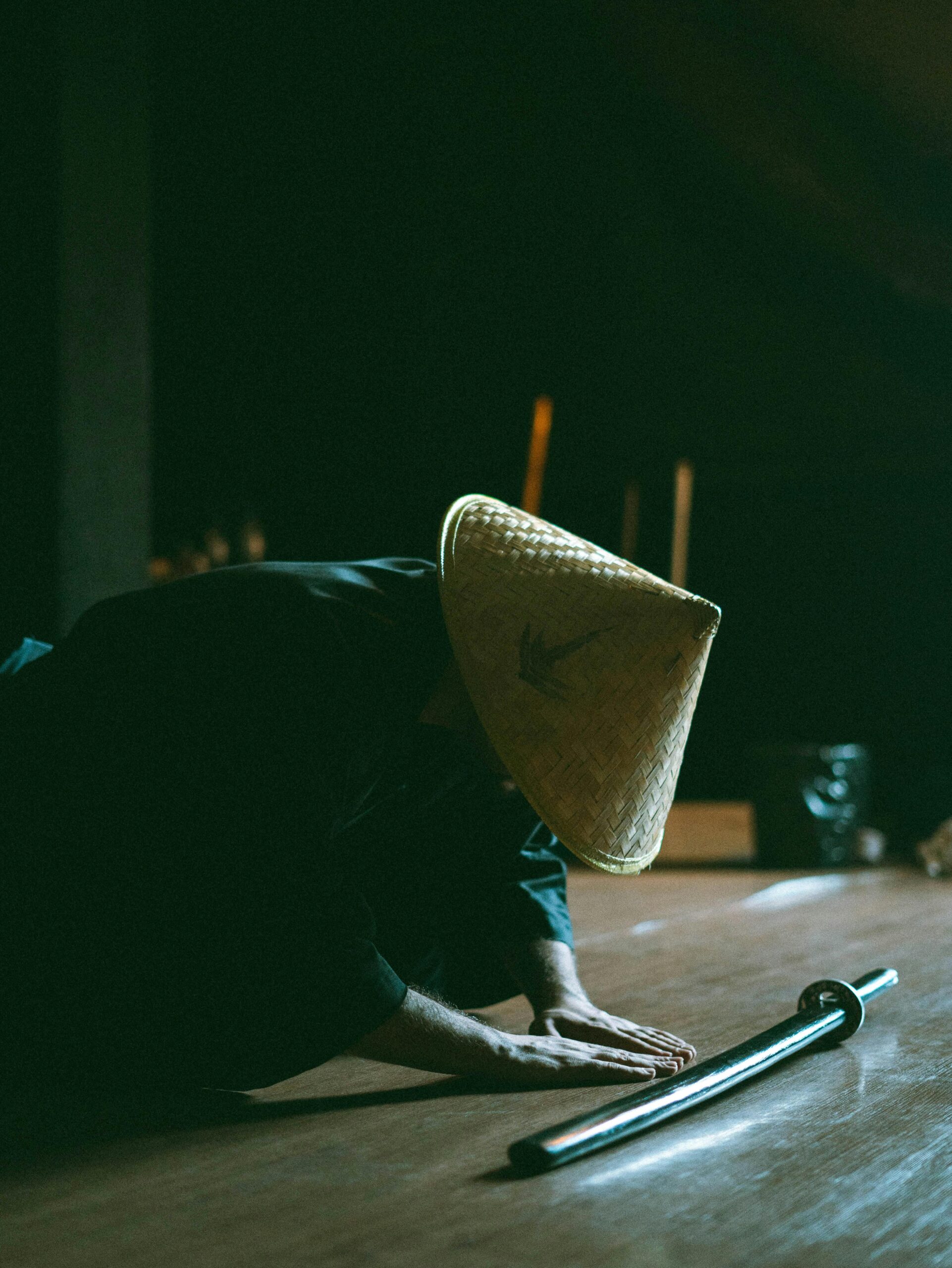 Person in a straw hat bowing deeply on the floor in front of a sword, evoking traditional Japanese respect and discipline