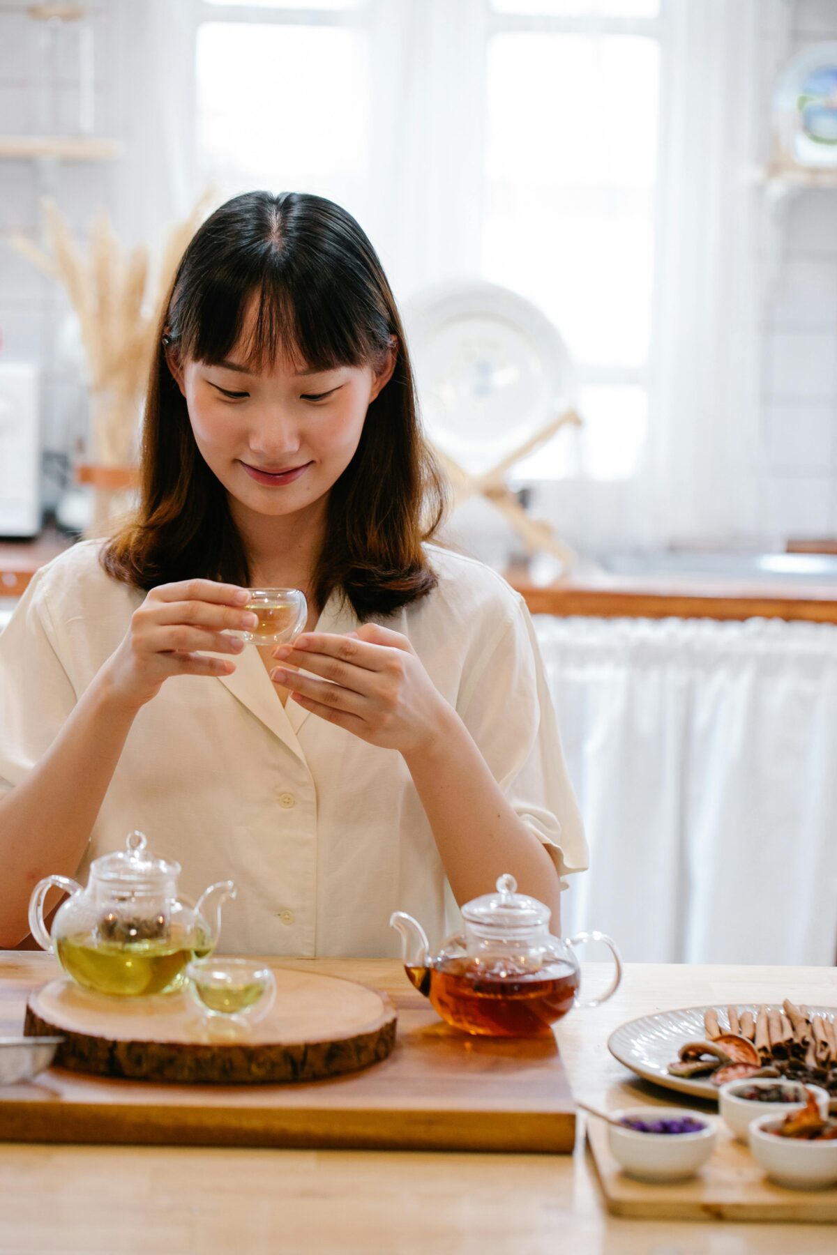 A young woman smiling softly while holding a small glass teacup, with green and black tea pots on the table in front of her.