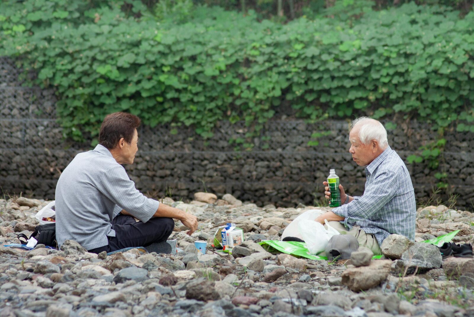 Two elderly men sitting outdoors, chatting and drinking tea by the river, symbolizing friendship and connection among Japan’s senior community