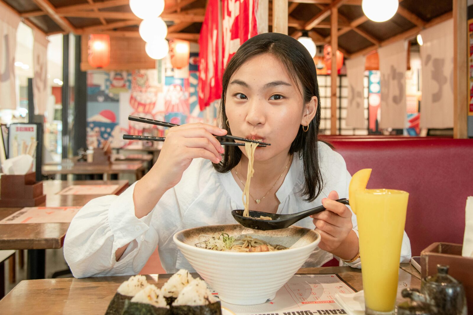 A woman happily eating ramen with chopsticks at a Japanese restaurant, with onigiri and a drink beside her.