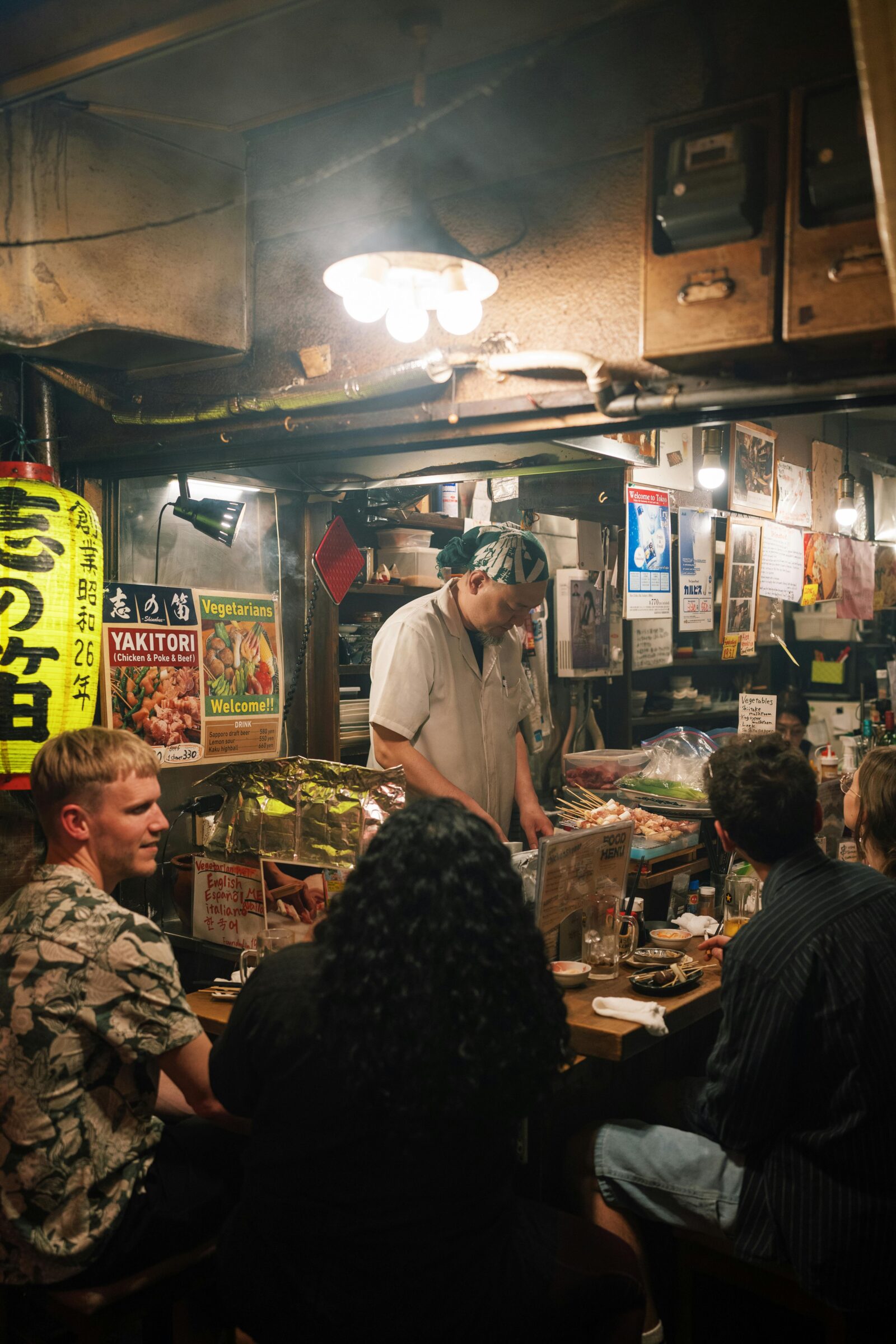 A yakitori chef grilling skewers inside a cozy stall, while customers enjoy food and drinks.