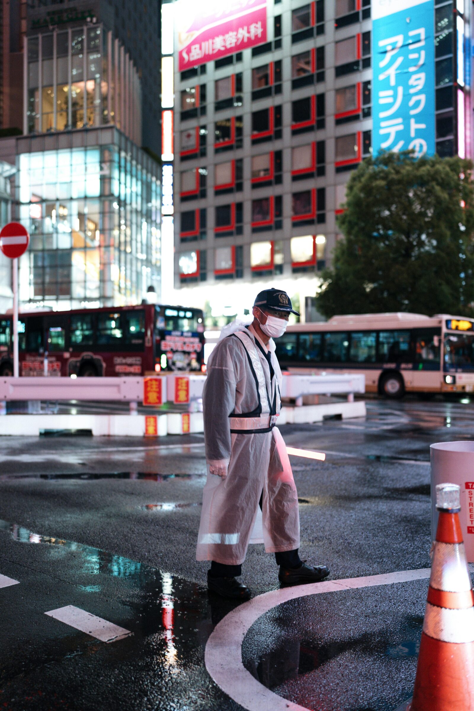 Masked traffic security guard in raincoat standing on wet Shibuya street at night with neon signs glowing in the background.
