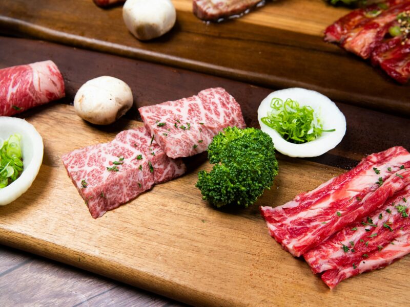 Slices of wagyu beef with mushrooms, broccoli, and onions on a wooden board, ready for Japanese yakiniku grilling.