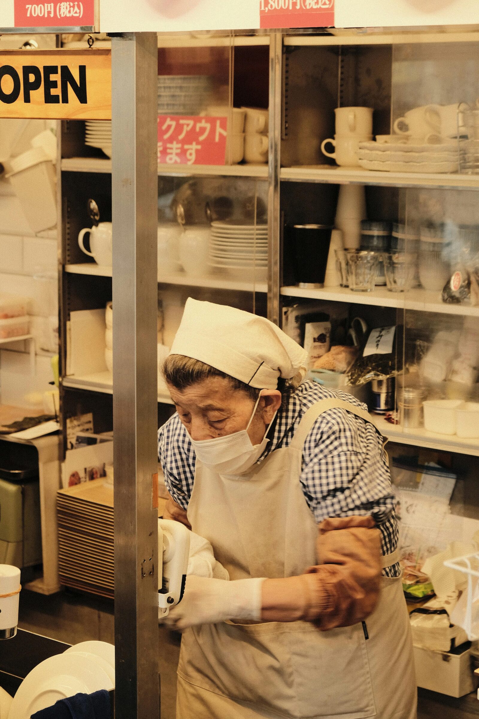 Older woman wearing a mask and apron working inside a Japanese kitchen, showing dedication and care.