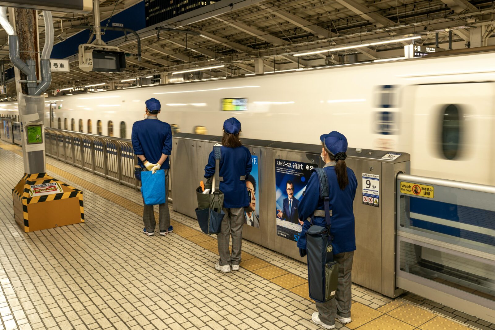 A team of uniformed Shinkansen cleaning staff standing neatly on the platform as a bullet train departs, reflecting Japan’s discipline, teamwork, and pride in service.