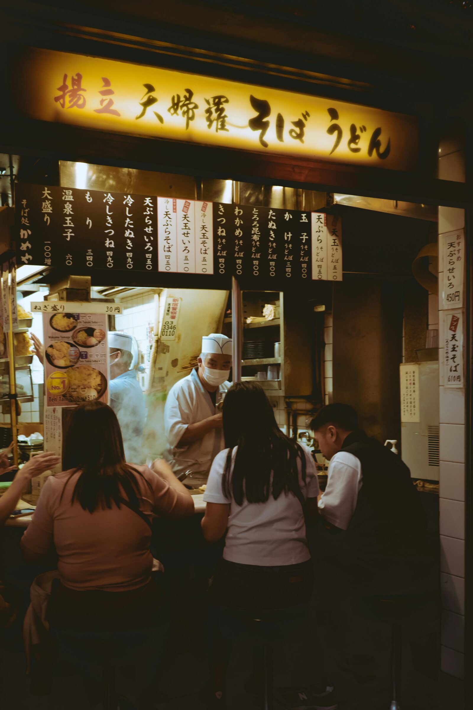 A small soba and udon shop in Japan, where a chef wearing a white uniform and mask serves steaming bowls of noodles to customers seated at the counter, capturing the warmth and intimacy of local dining culture.