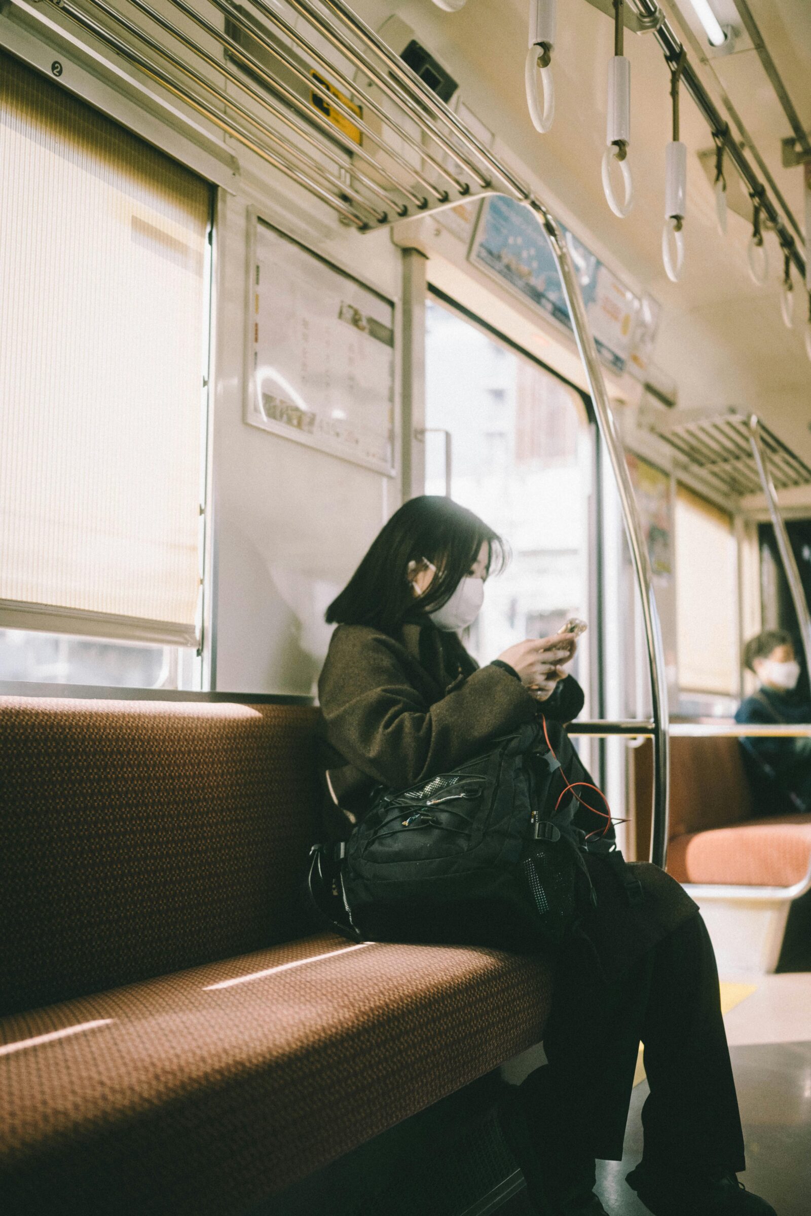 Young woman in a brown coat wearing a mask, sitting alone on a train seat and looking at her smartphone.