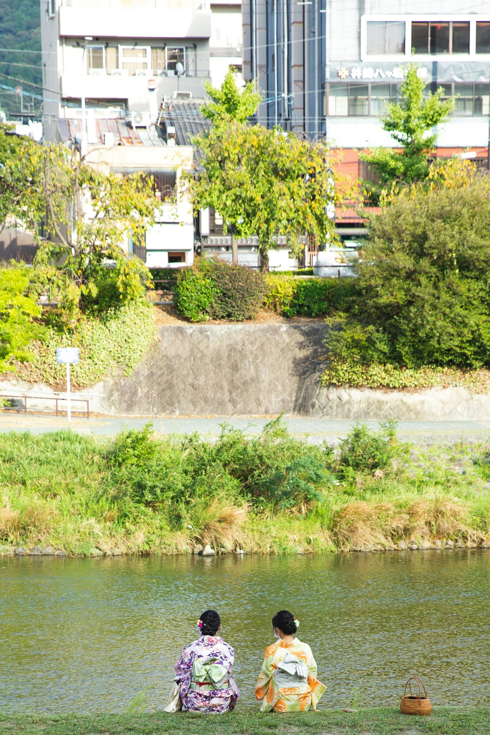 Two women in colorful kimono sit quietly by the riverbank, enjoying a peaceful afternoon in a Japanese town.
