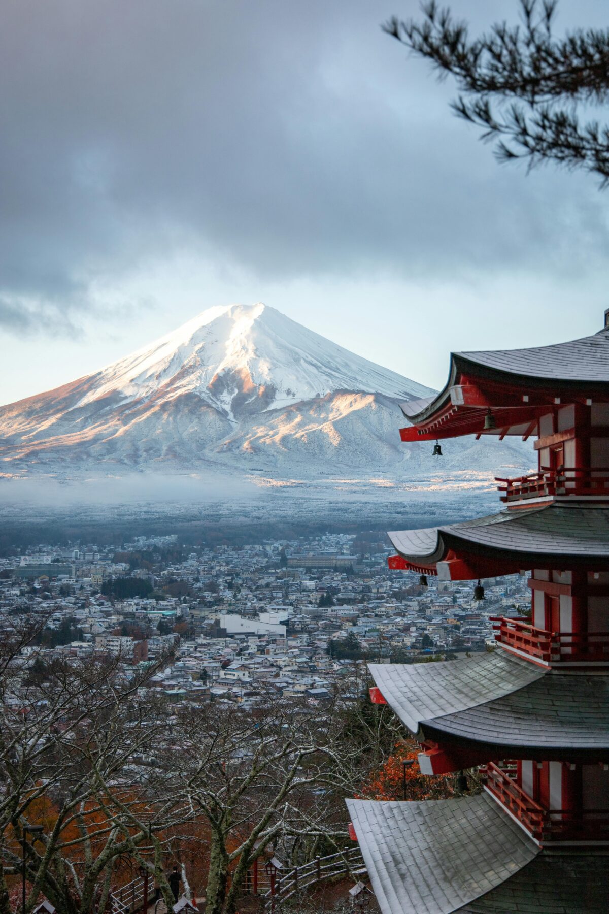 A stunning view of Mount Fuji with snow-covered peak seen from Chureito Pagoda in Japan, framed by the red pagoda roof and a winter cityscape below.