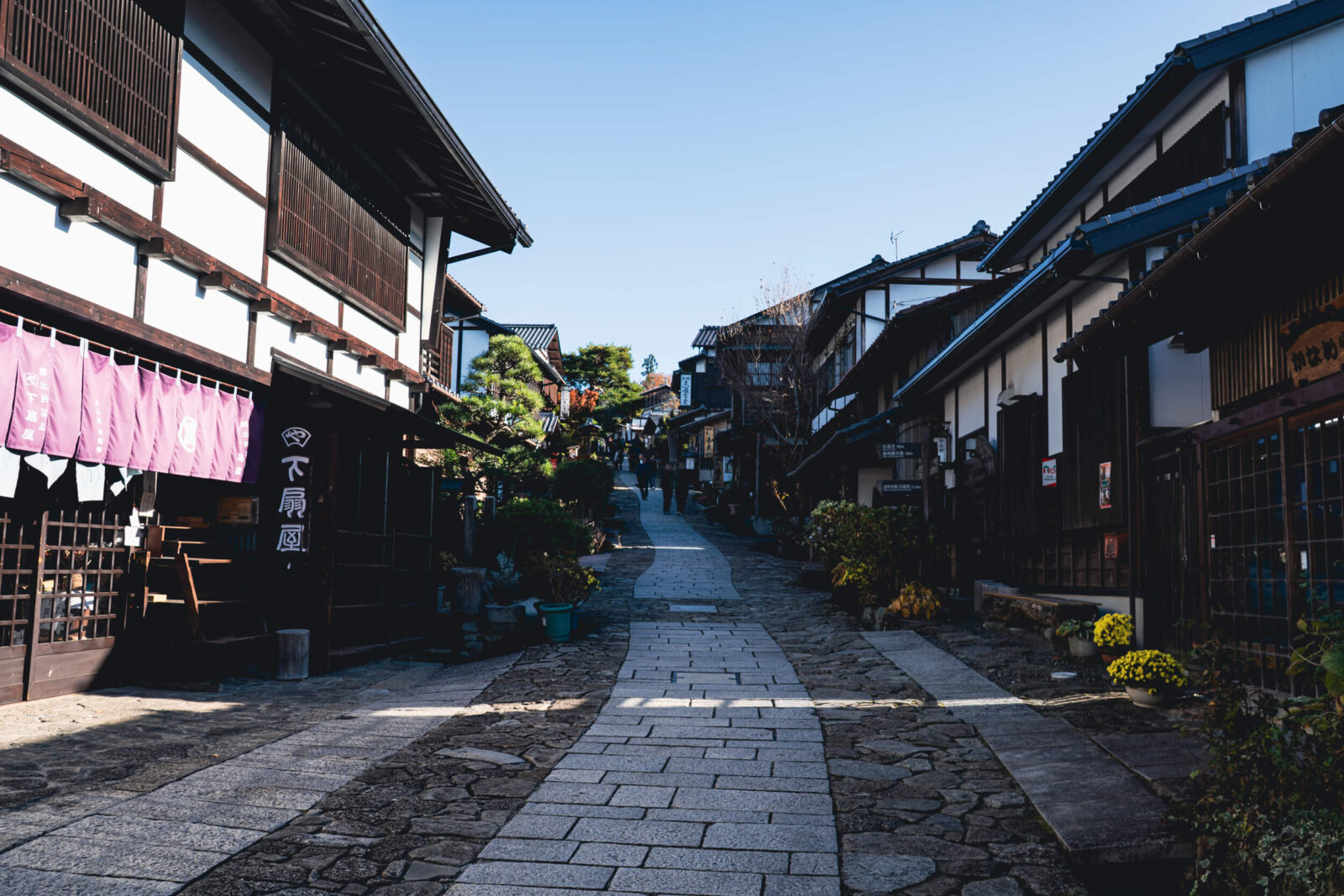 A scenic downhill street in Magome-juku with traditional wooden houses, autumn foliage, and visitors walking under clear mountain skies.
