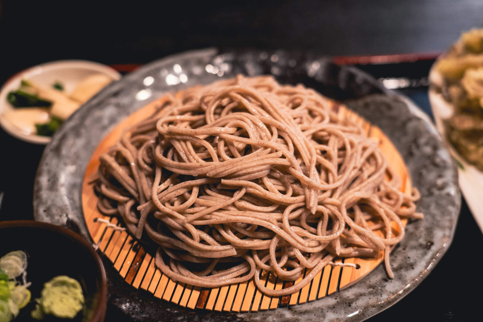 A plate of chilled zaru soba noodles arranged on a bamboo mat, served with dipping sauce and condiments.
